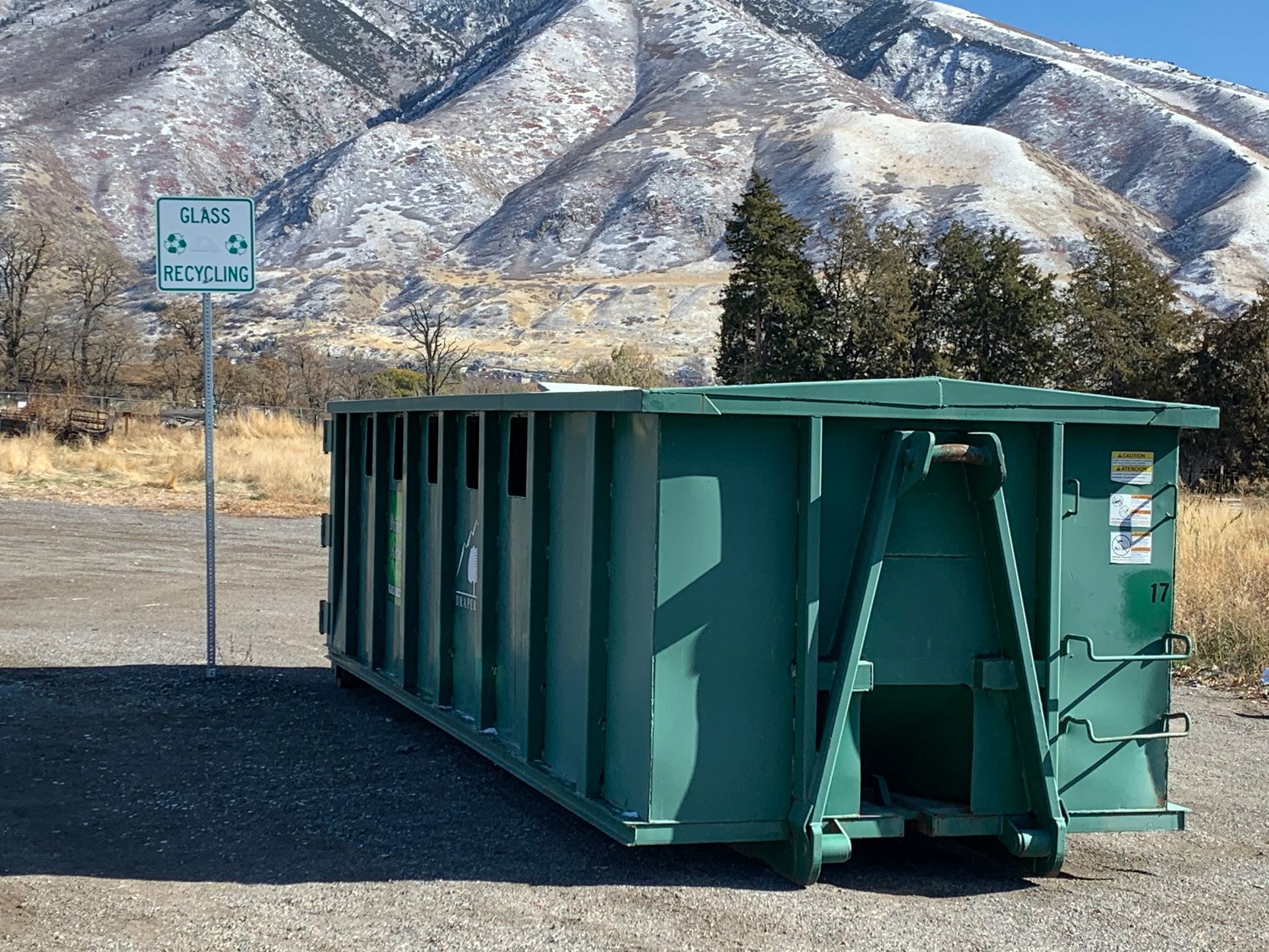 A large green recycling dumpster sits on a gravel lot in front of snow-capped mountains.