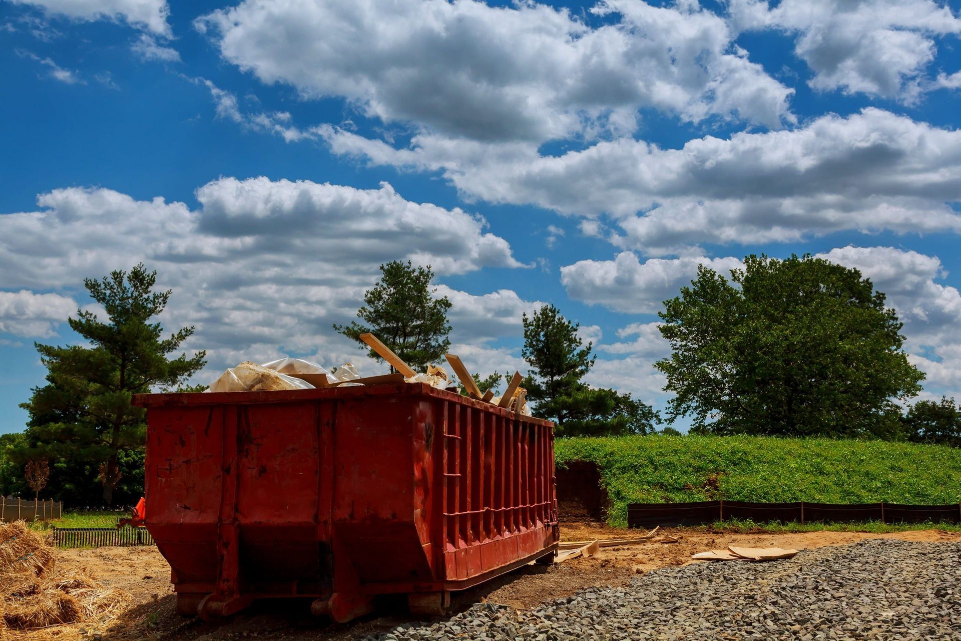 A red construction dumpster filled with debris sits on a gravel lot under a bright blue, partly cloudy sky.