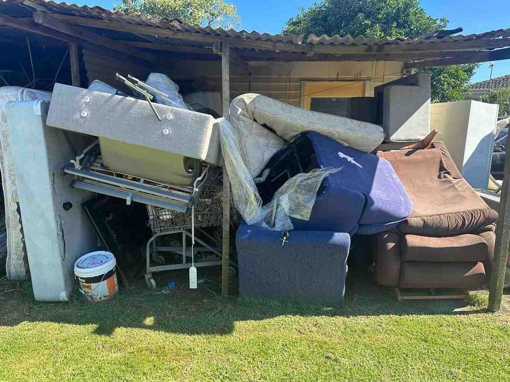 A cluttered outdoor storage area filled with old mattresses, furniture, and a paint bucket under a corrugated metal roof.
