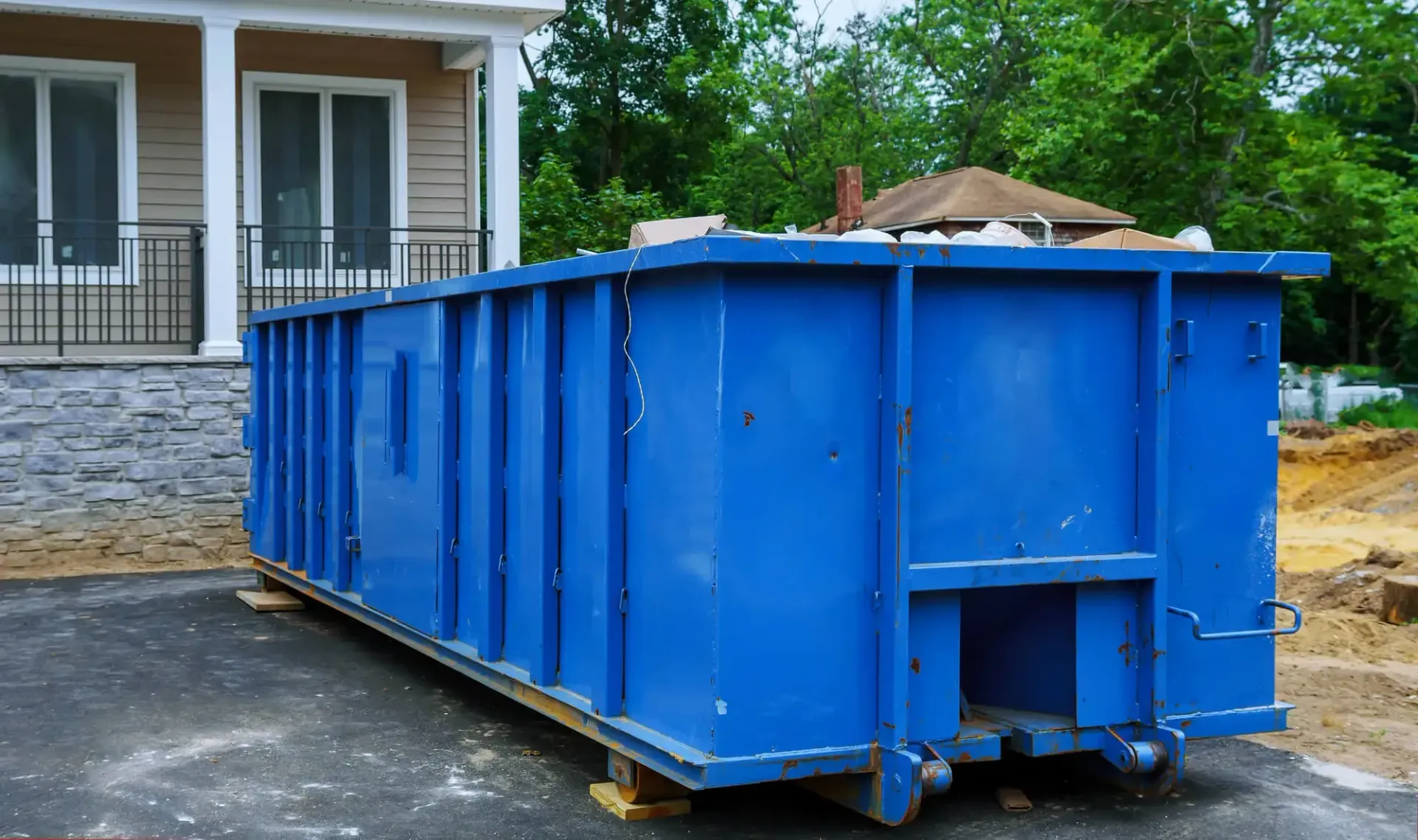 A large, rectangular, bright blue metal dumpster sits on an asphalt driveway next to a residential house.
