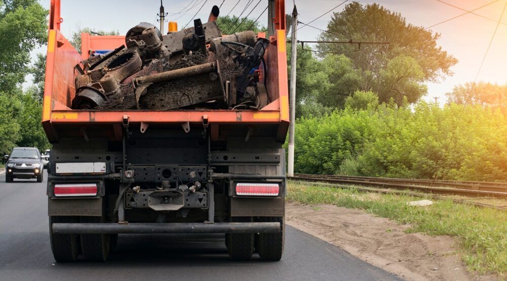 An orange dump truck filled with scrap metal travels down a paved road beside railroad tracks.