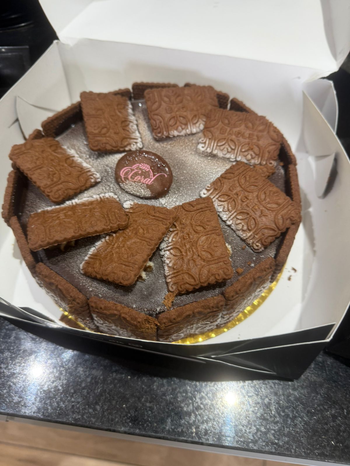 Pastel de chocolate con galletas alrededor del borde, espolvoreado con azúcar en polvo, en una caja abierta.