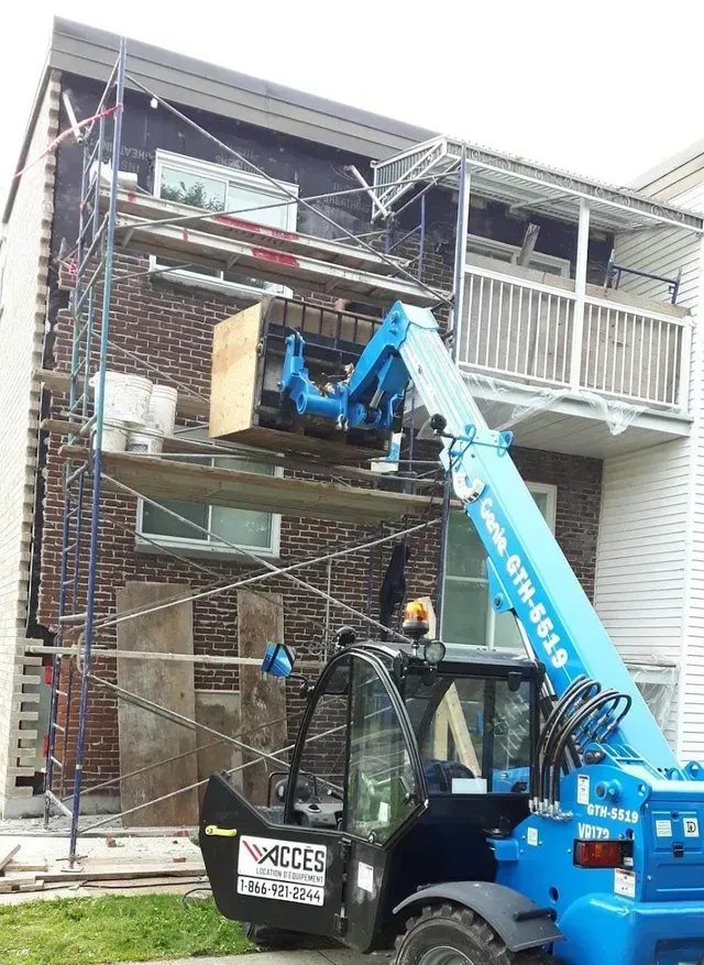 Chariot élévateur bleu soulevant une boîte en bois près d'un bâtiment en briques avec un échafaudage.