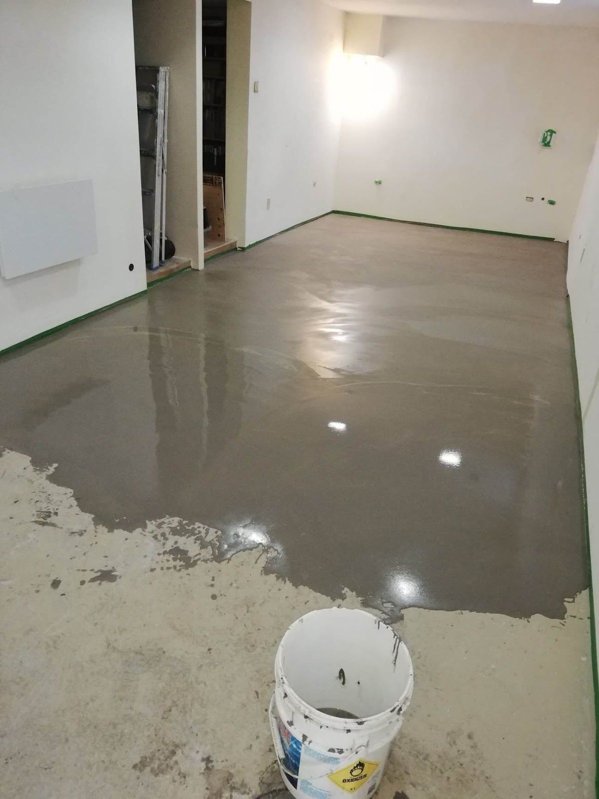 Sol en béton gris fraîchement coulé dans une pièce avec des murs blancs, du ruban adhésif vert et un seau.