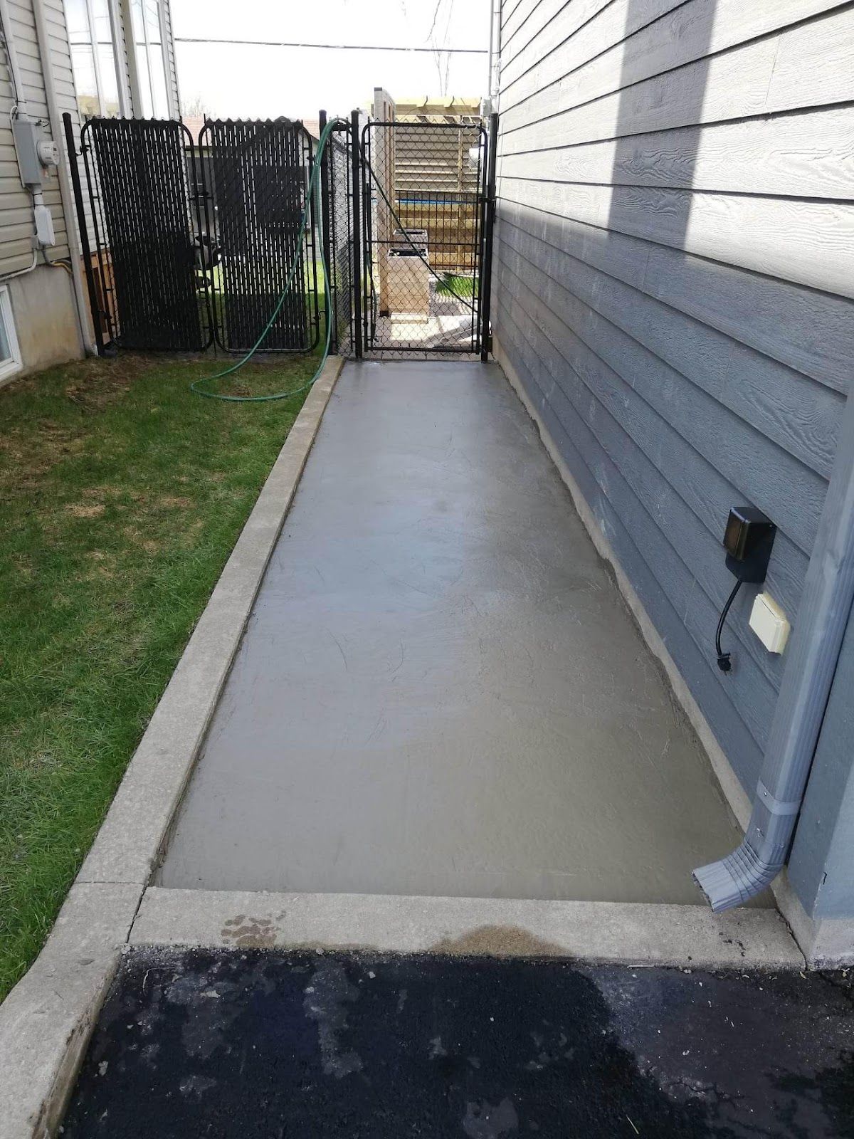 Allée en béton longeant une maison grise, menant à un portail. Elle comprend de la pelouse, une gouttière et une prise électrique.