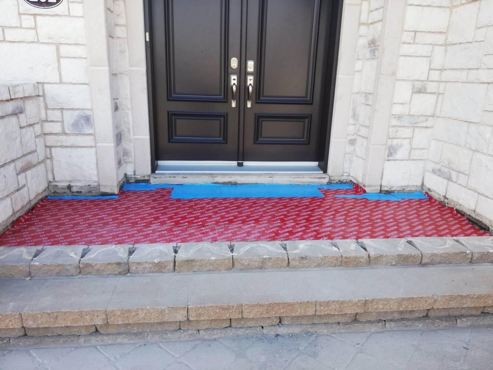 Un tapis rouge recouvre l'entrée en béton, menant à une double porte noire. Des marches en pierre grise se trouvent en contrebas.
