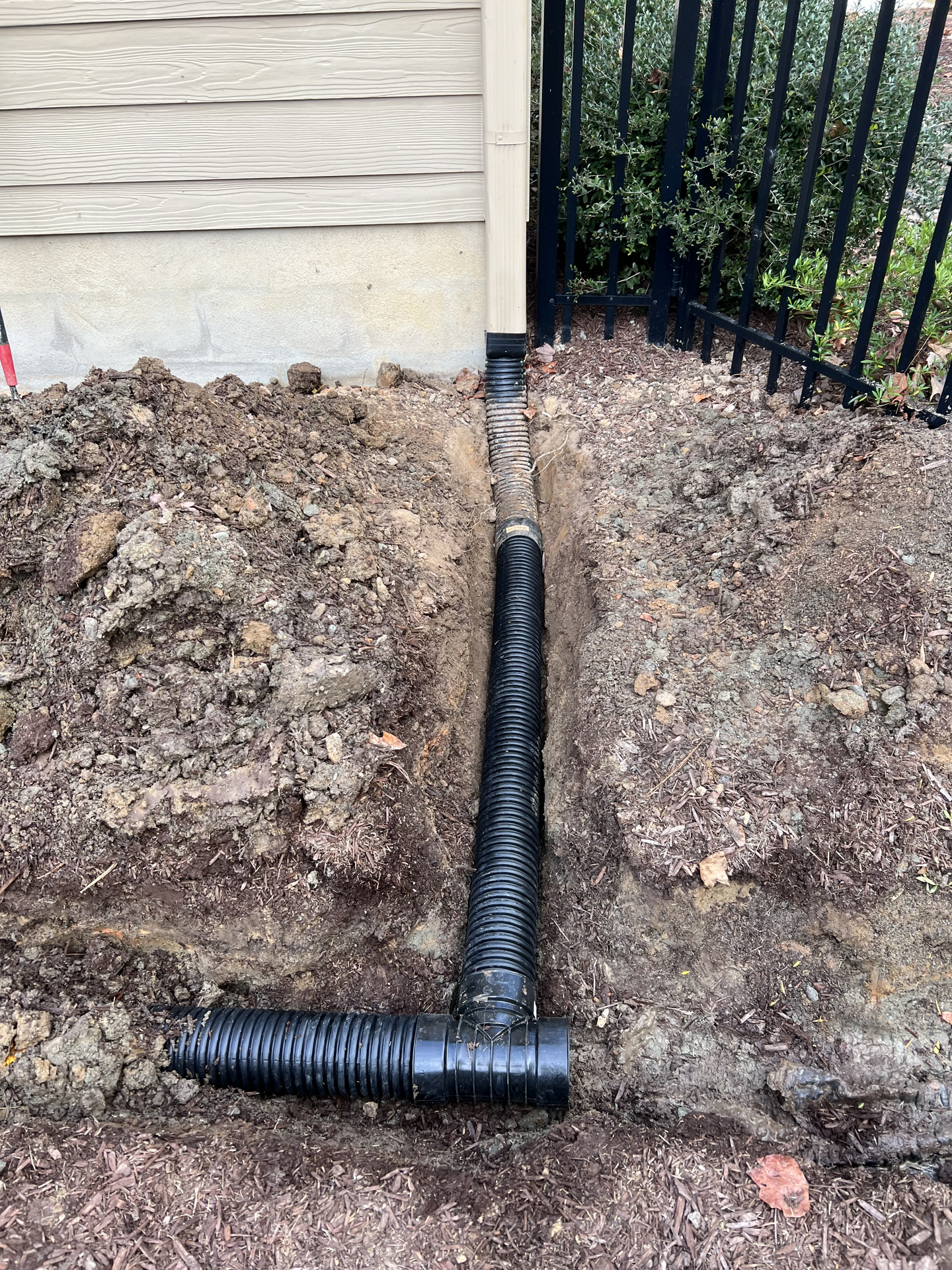 Black drainage pipe in a trench next to a building and fence, surrounded by dirt.