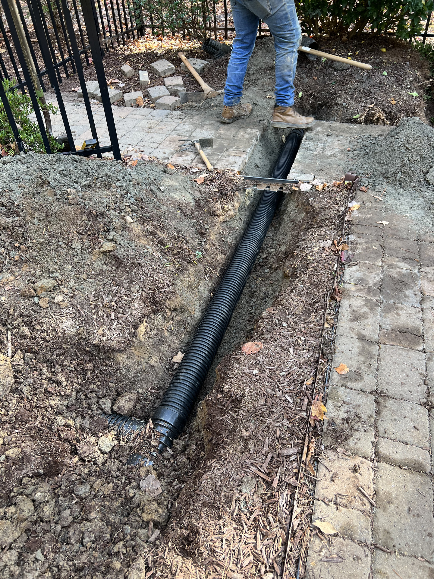 Person installing black corrugated drainage pipe in a trench next to a brick path.