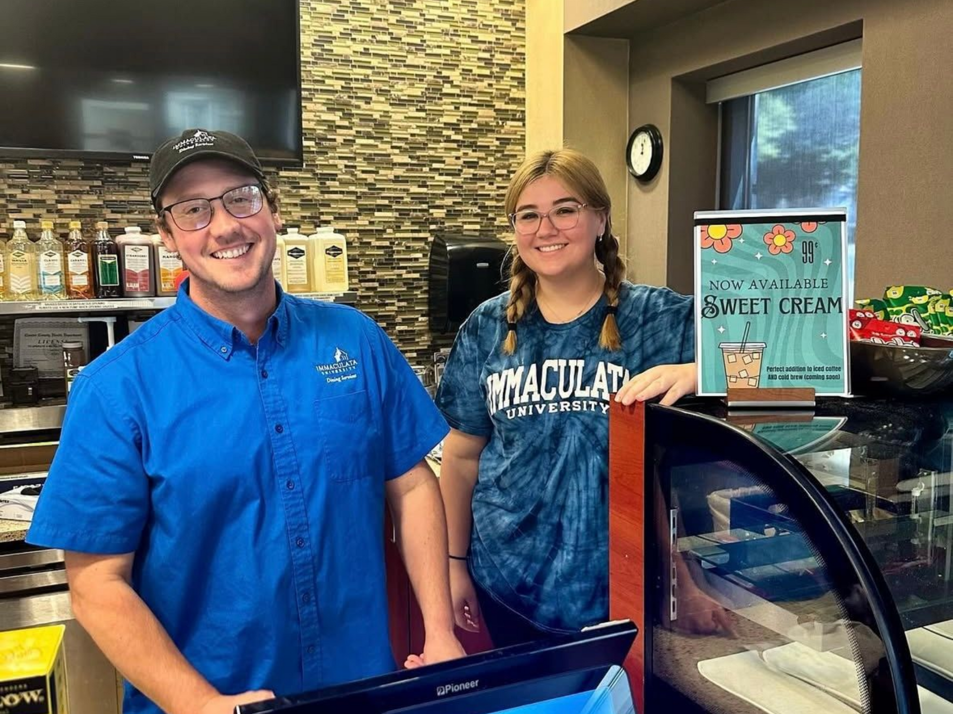 Two people smiling behind a coffee shop counter. One in blue, one in tie-dye, next to a 