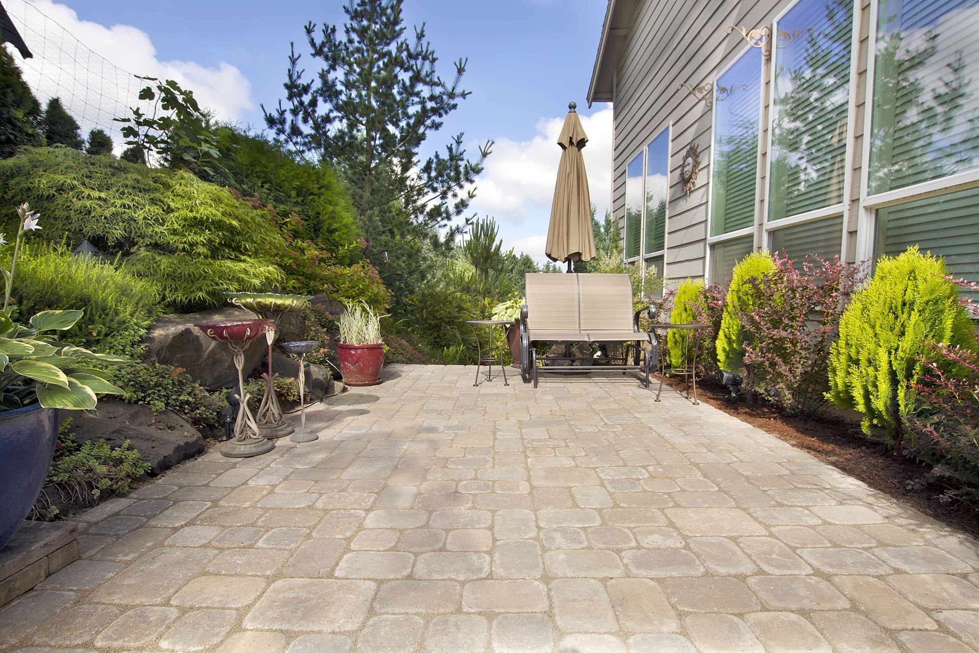 Patio with stone pavers, bench, and umbrella next to a house and garden under a sunny sky.