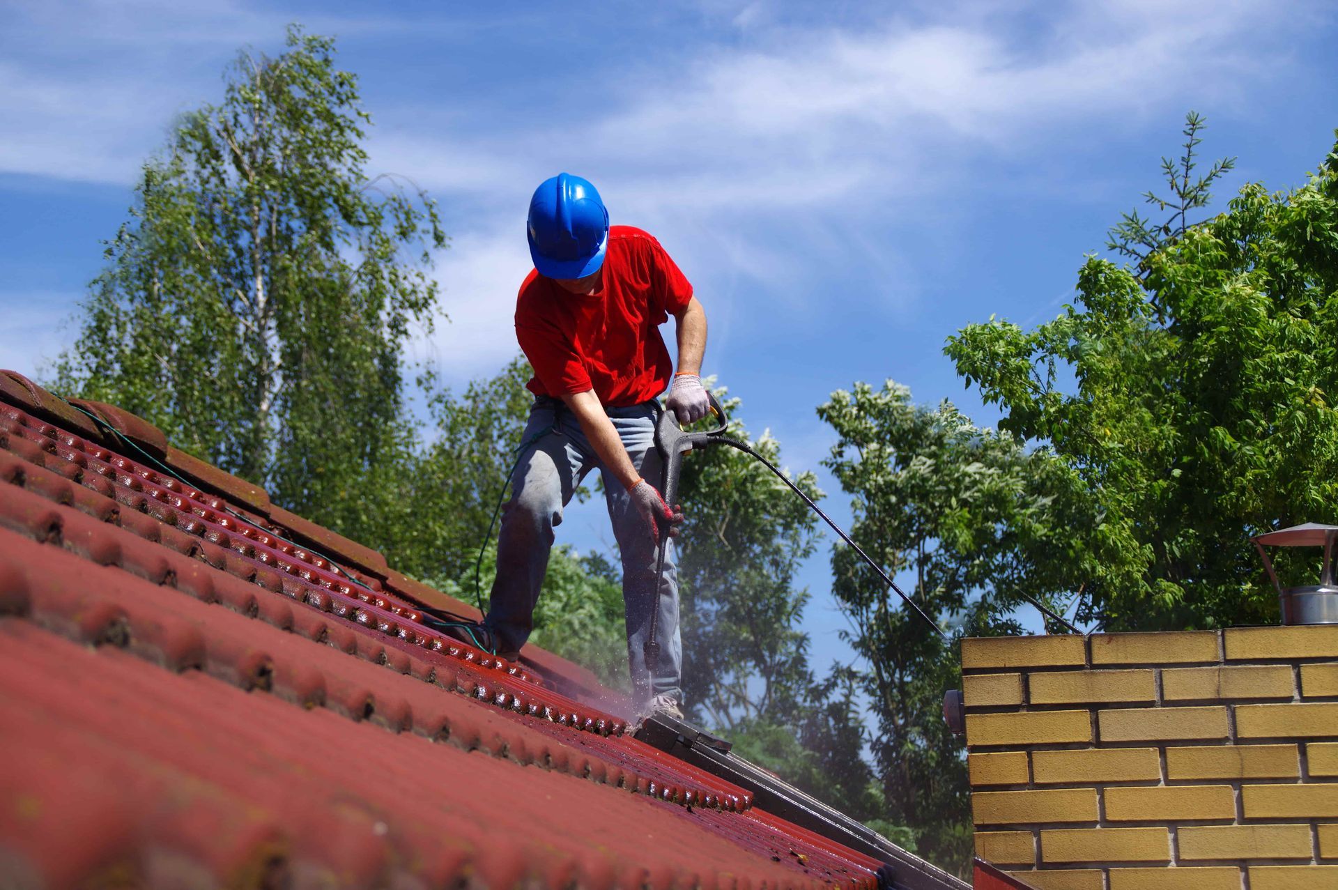 Man in blue helmet power washing a red tile roof on a sunny day.
