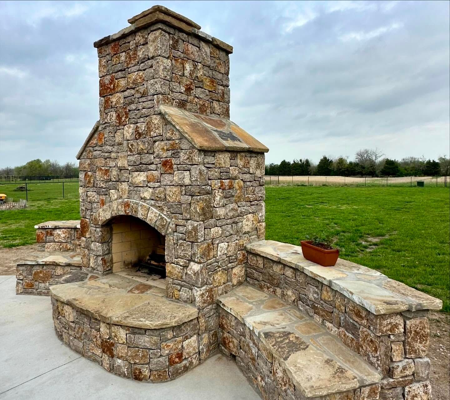 Stone outdoor fireplace with chimney, built-in seating, set in a grassy field under a cloudy sky.