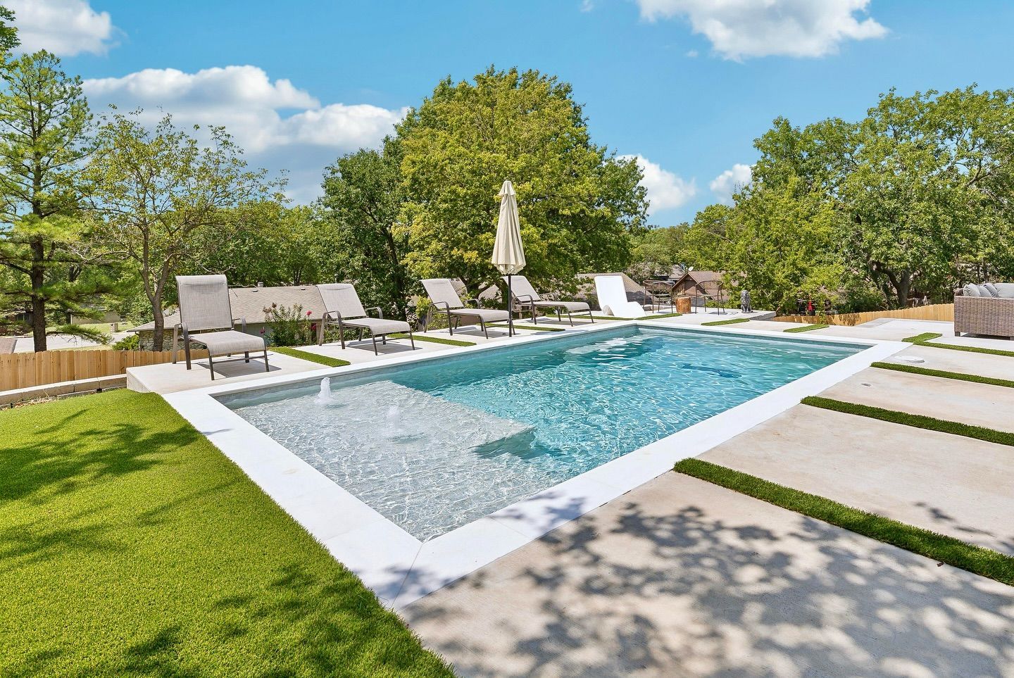 Rectangular pool surrounded by concrete and green grass; lounge chairs, trees, and blue sky.