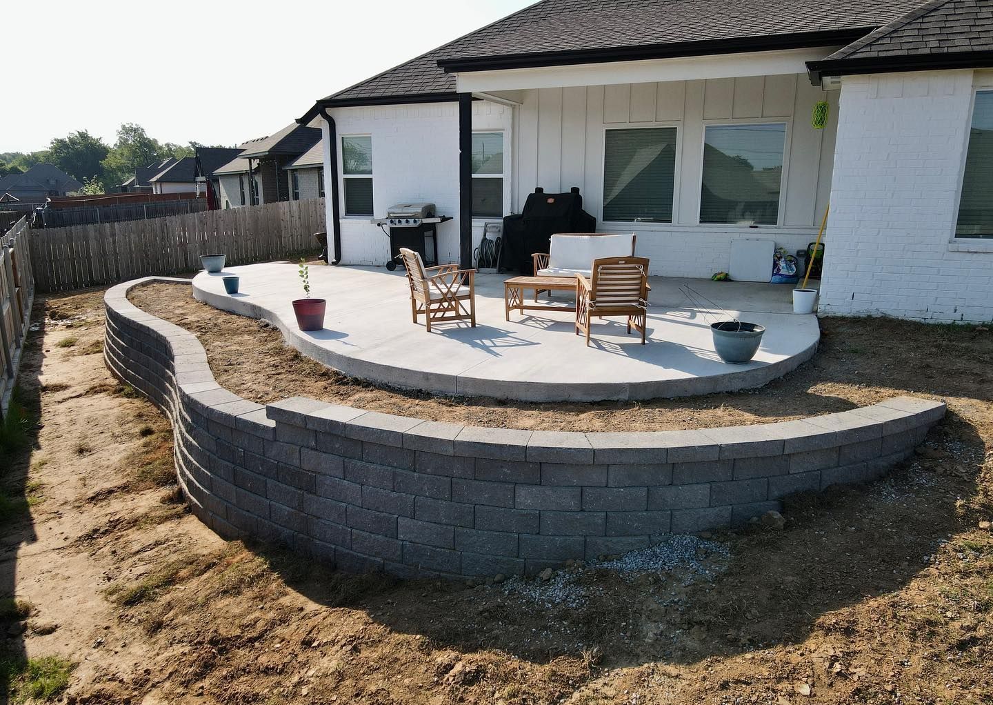Gray block retaining wall curves around a concrete patio with outdoor furniture, near a house.