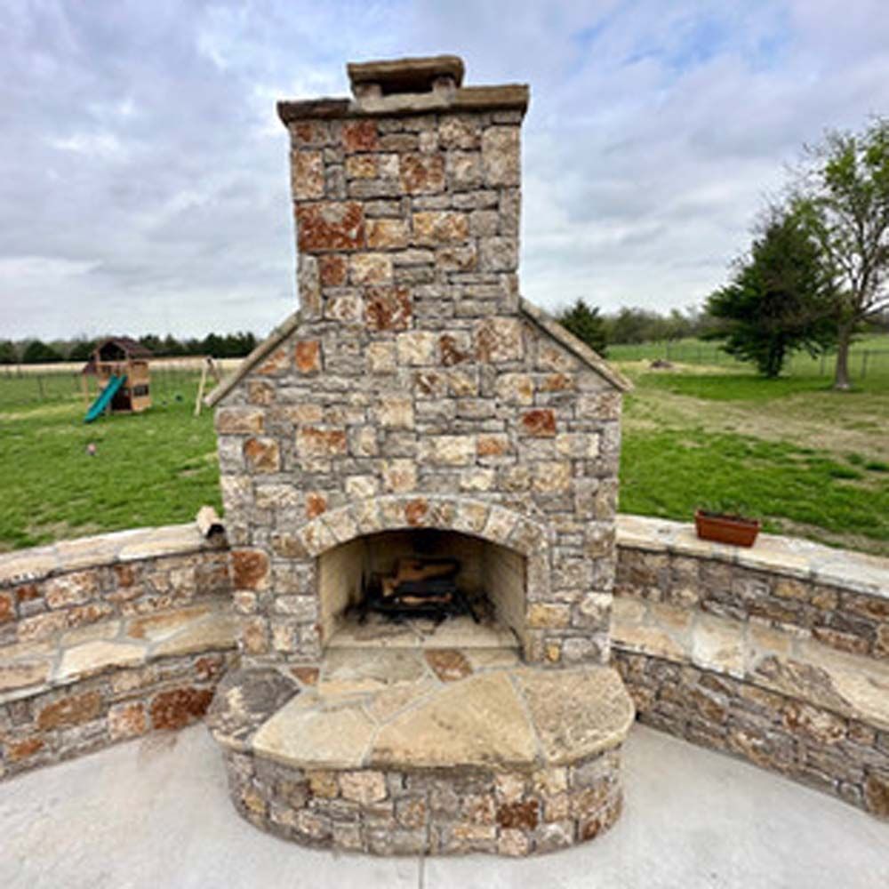 Stone outdoor fireplace with curved hearth, flanked by low walls on a concrete patio, set in a grassy field.