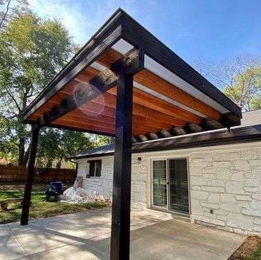 A wooden pergola with a black frame and natural wood interior, attached to a white stone house.