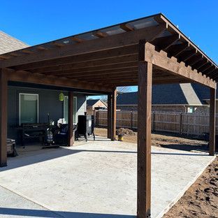 Wooden pergola over a concrete patio, attached to a house. Dark wood, blue sky.