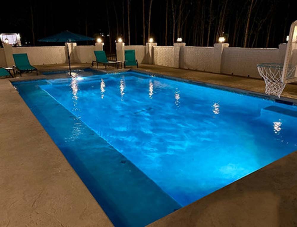 A nighttime shot of a blue-lit pool with lounge chairs, basketball hoop, and surrounding white walls.