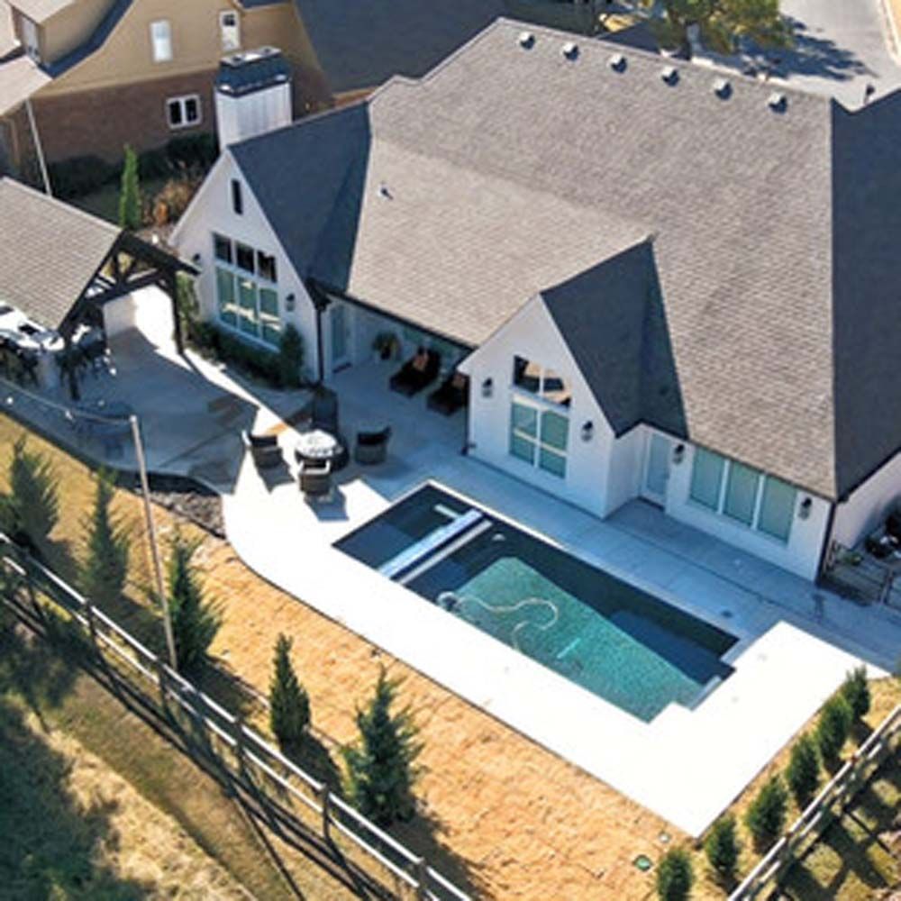 Aerial view of a modern house with a pool and patio. White exterior, dark roof, green yard, and fence.