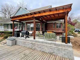 Wooden pergola over a patio with seating, grill, and a brick house in the background.