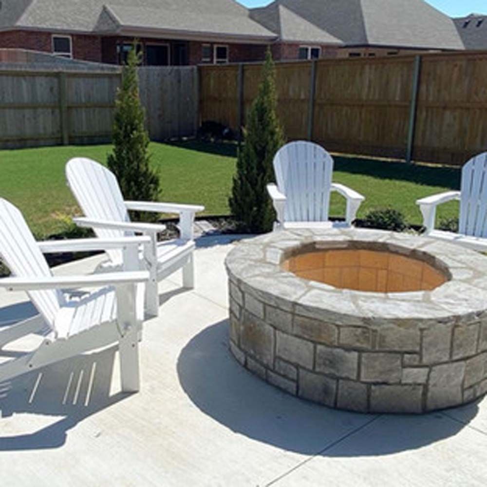 Four white Adirondack chairs surround a stone fire pit on a concrete patio in a backyard.