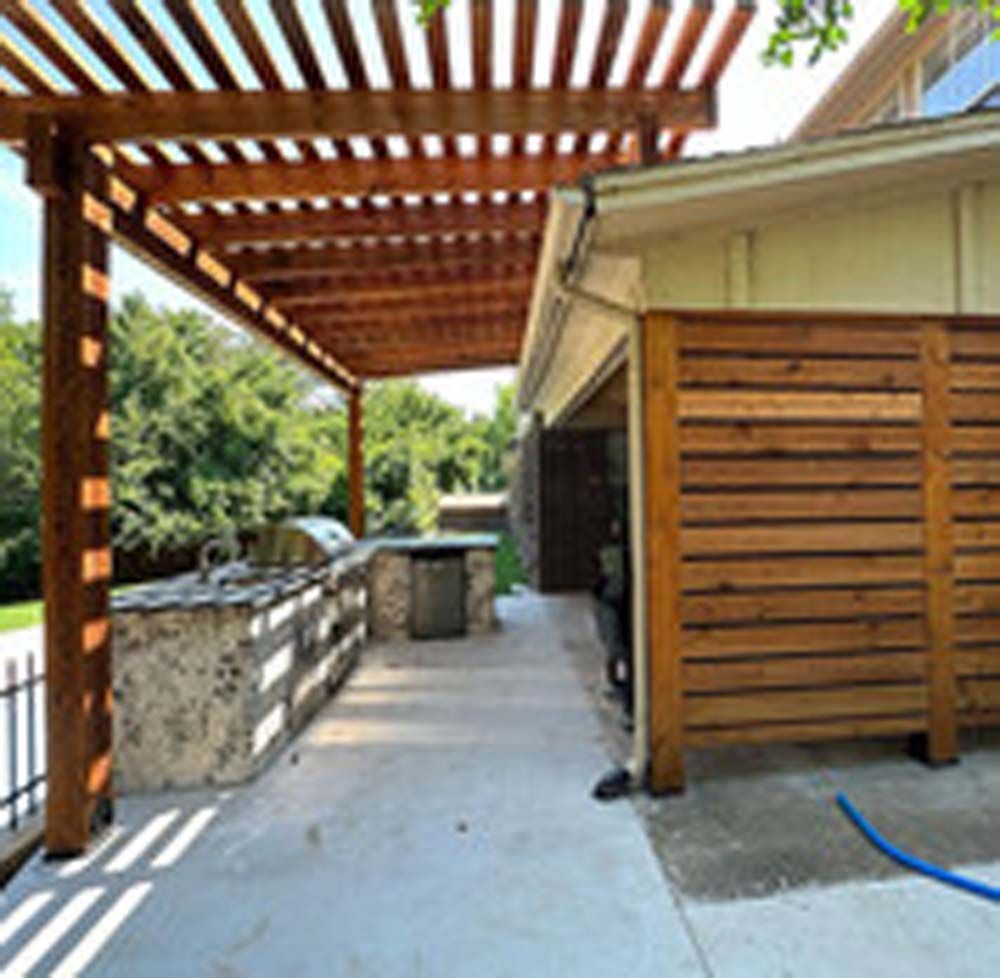 Outdoor kitchen with pergola, concrete counter, grill, and horizontal wood fence.