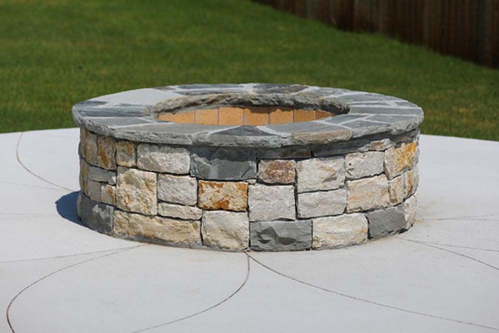 Circular stone fire pit on a concrete patio with a green lawn background.