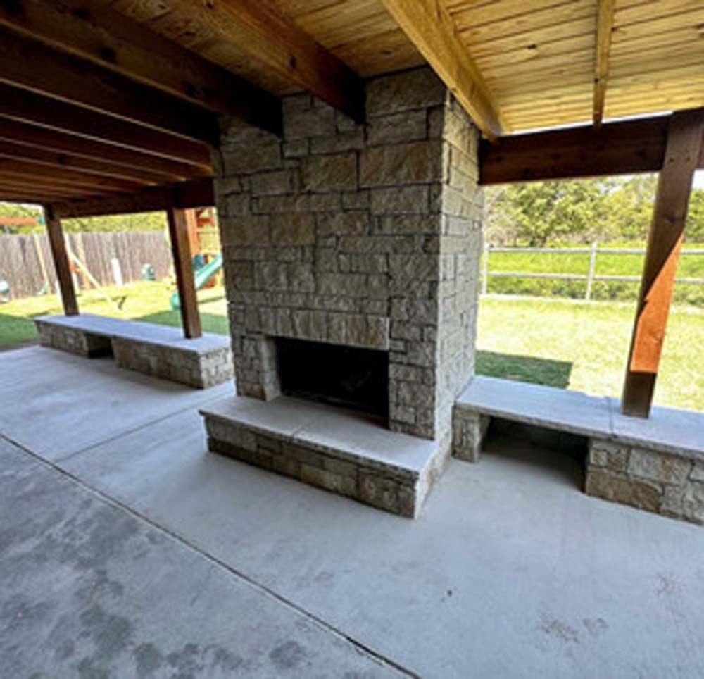 Stone fireplace with benches on patio under a wood-beamed awning; backyard in view.