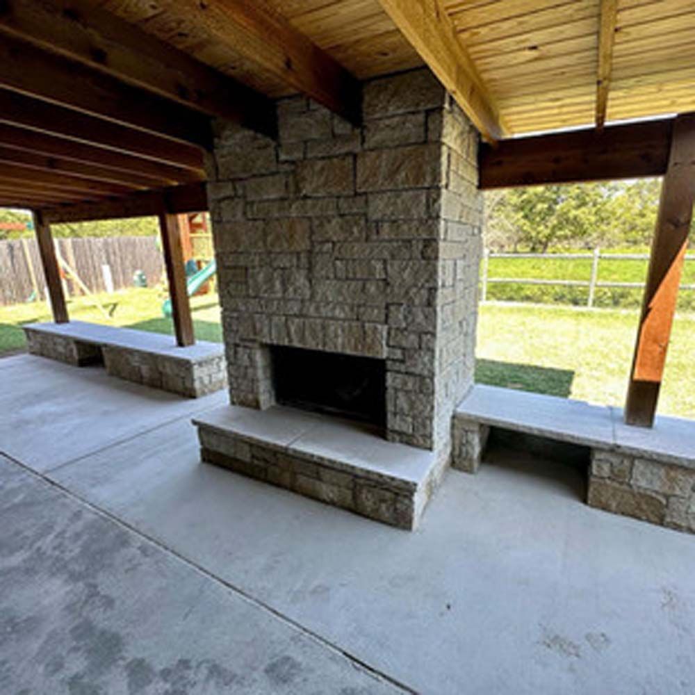 Outdoor fireplace made of stone, with matching stone benches on concrete patio, under a wooden pergola.