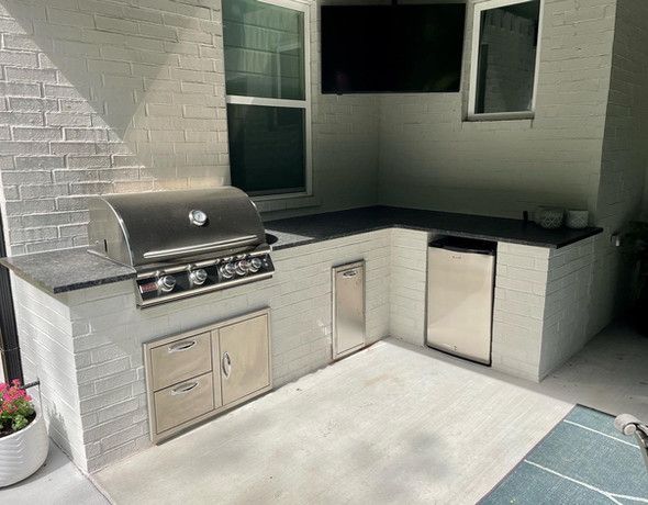 Outdoor kitchen with grill, refrigerator, and sink built into brick walls, beneath a TV.
