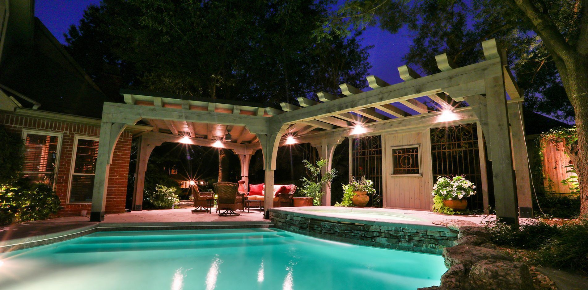 Pool at night with illuminated pergola, seating, and surrounding greenery.