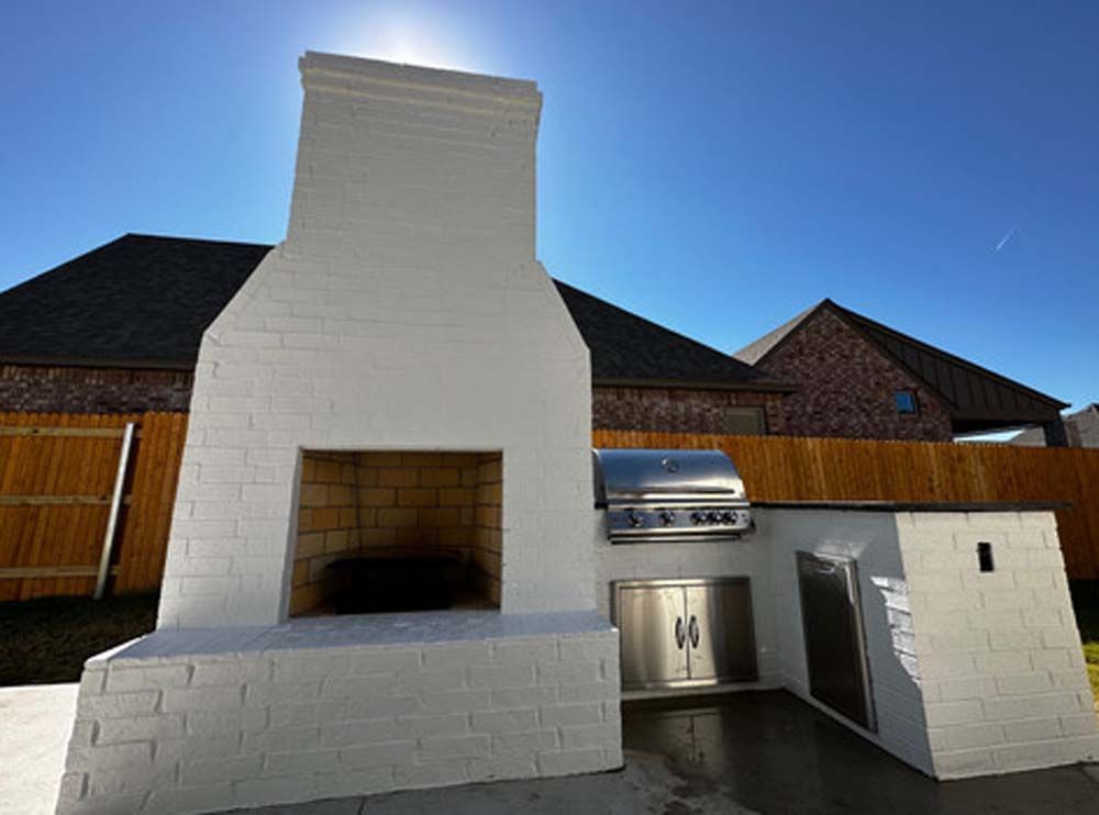 White brick outdoor kitchen with a fireplace, grill, and cabinets against a bright blue sky.