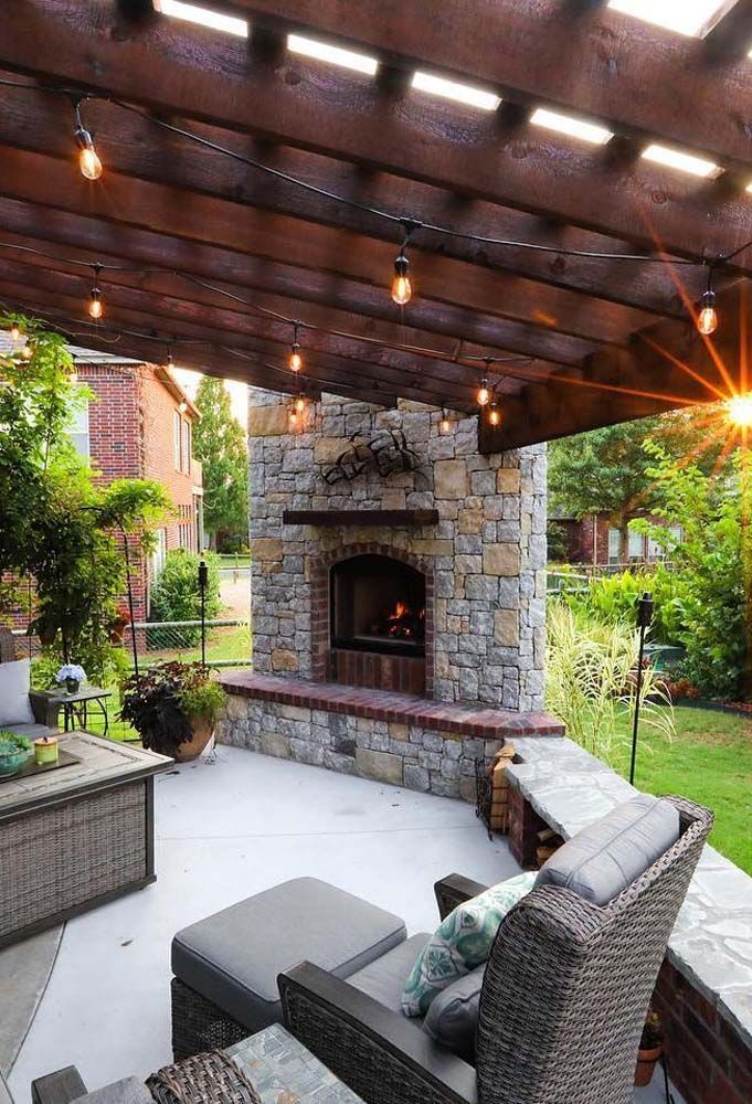 Patio with stone fireplace under wooden pergola, string lights, seating, and lush greenery.