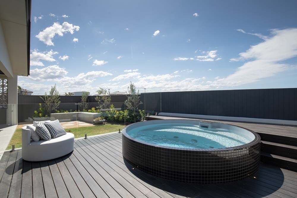 Backyard with a round hot tub on a deck, lounge chair, landscaping, and a fence under a blue sky.