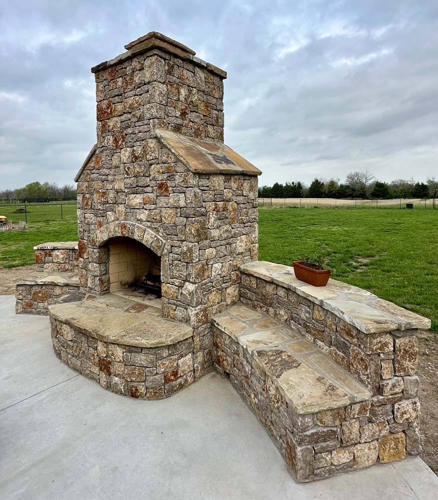 Stone outdoor fireplace with seating on a concrete patio, overlooking a grassy field under cloudy sky.
