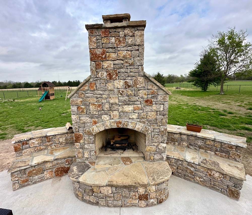 Stone outdoor fireplace with seating in a grassy yard under a cloudy sky.