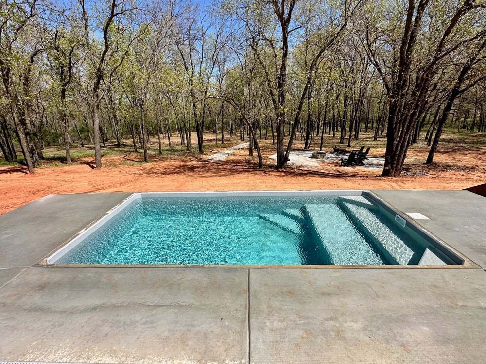 A rectangular pool with steps sits in a concrete patio, surrounded by a forest.