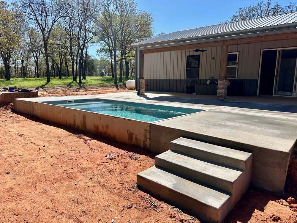 Concrete pool with steps, adjacent to a metal-sided building on red dirt. Blue water.