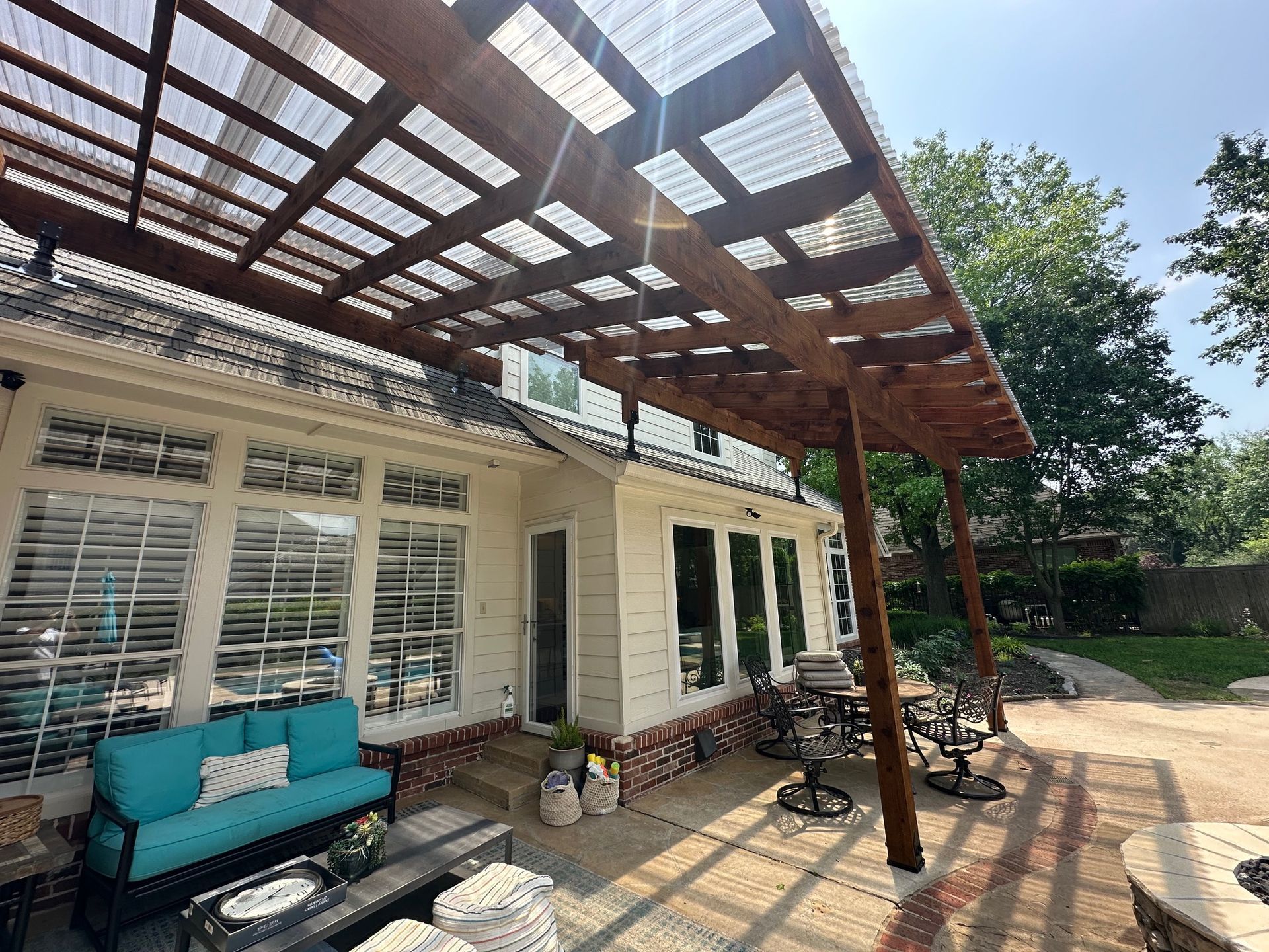 A pergola extends from a white house's brick patio; blue couch and chairs; clear roofing; sunny day.