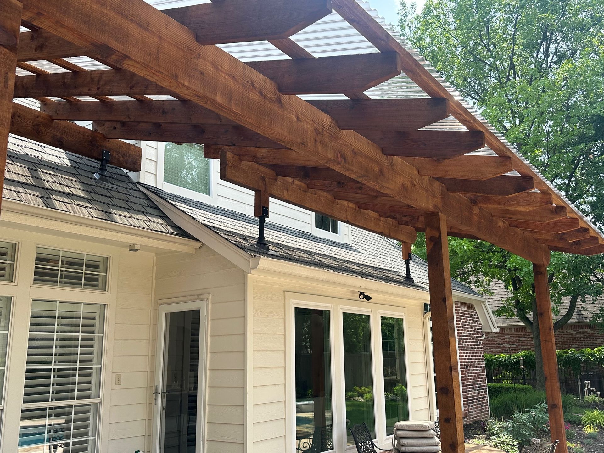 Wooden pergola attached to a light-colored house with windows.