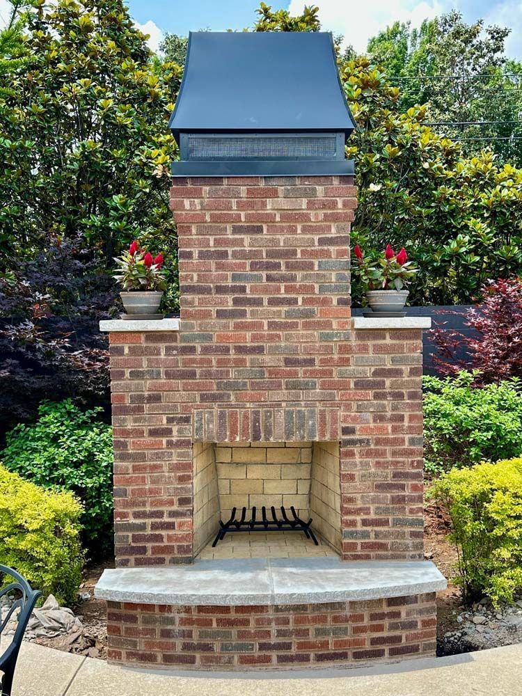 Brick outdoor fireplace with a black metal hood, surrounded by greenery.