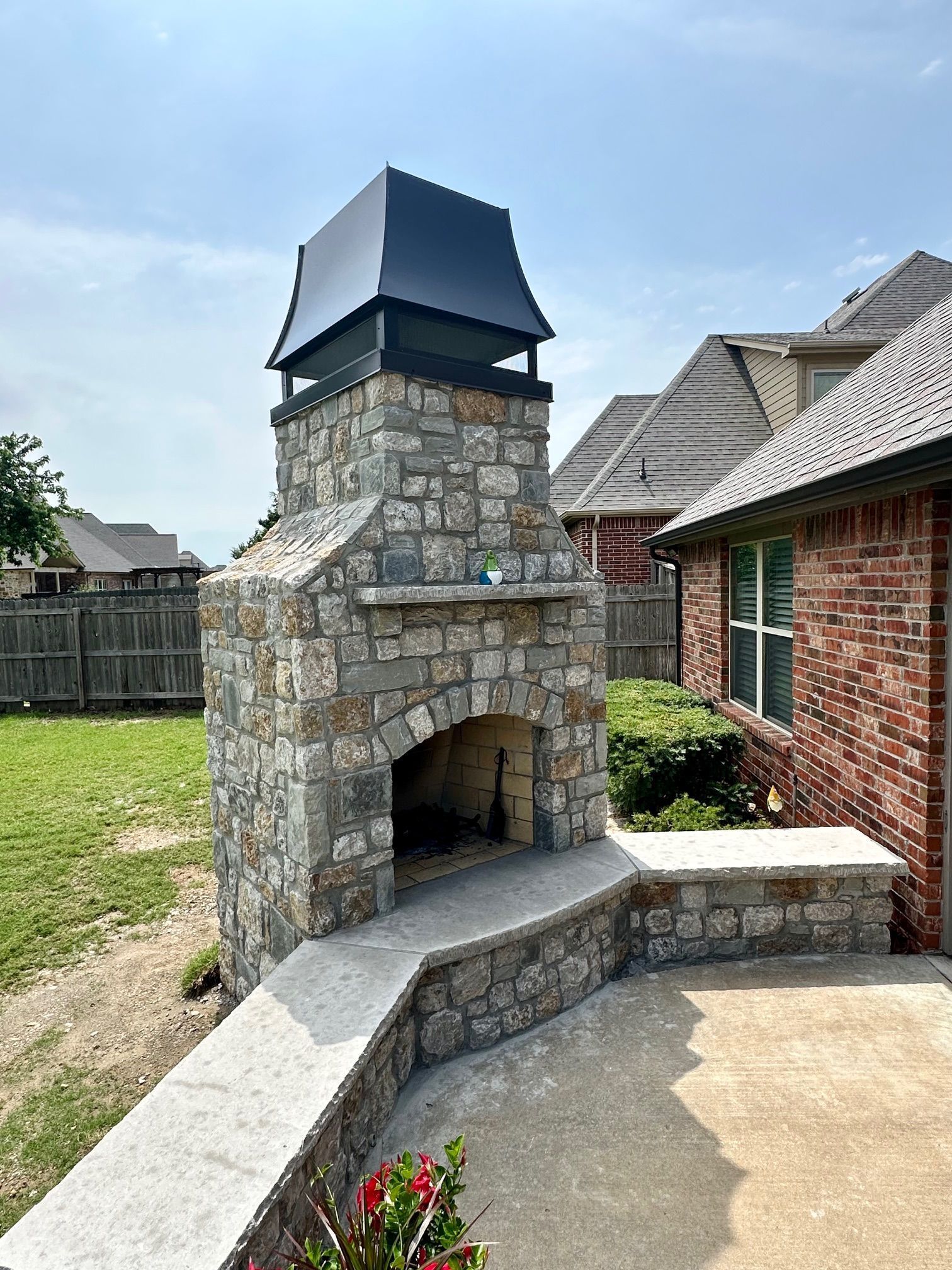 Outdoor stone fireplace with black metal cap, gray stone, on a patio.