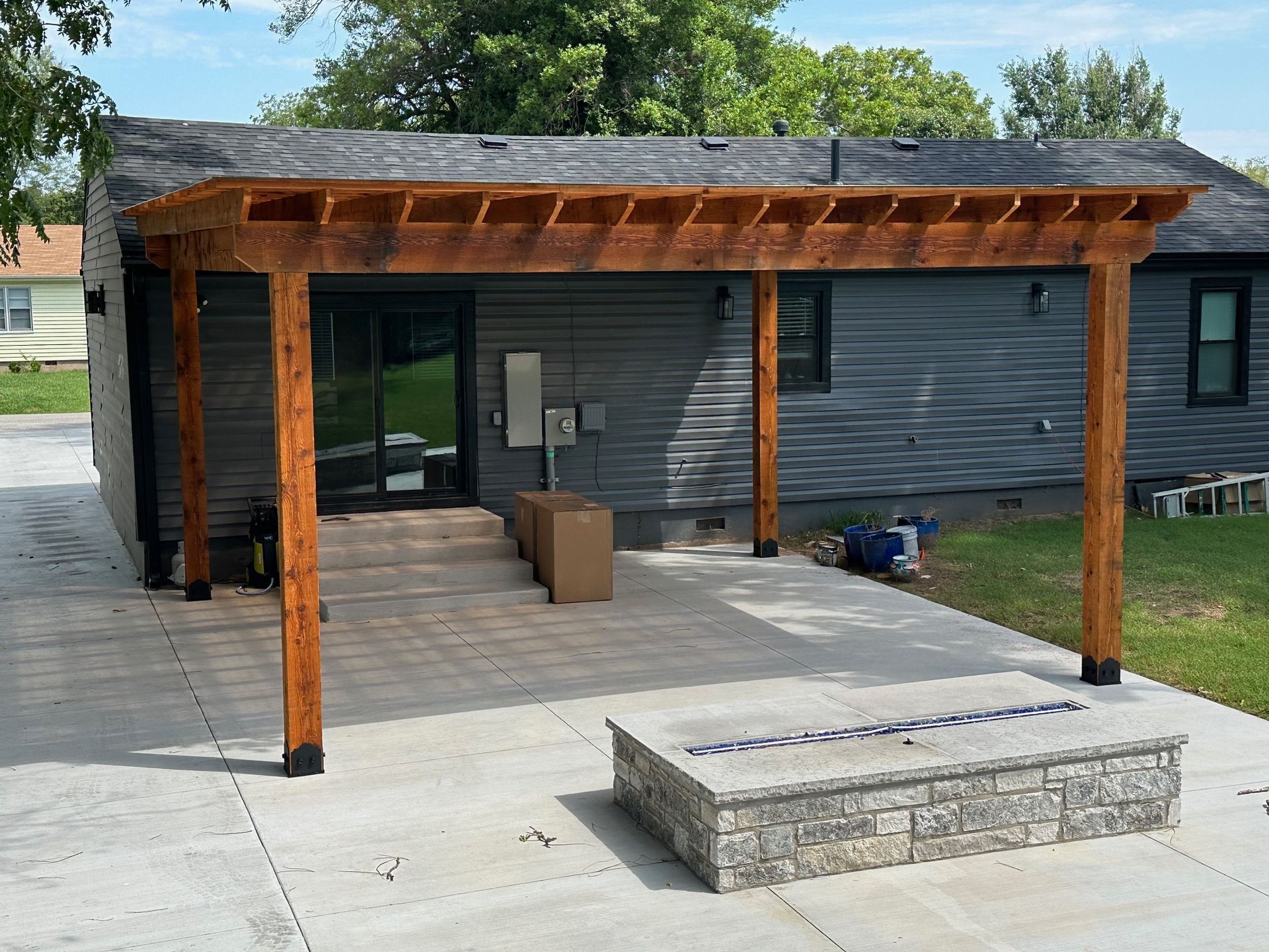 Wooden pergola over a concrete patio. Gray house with a fire pit.