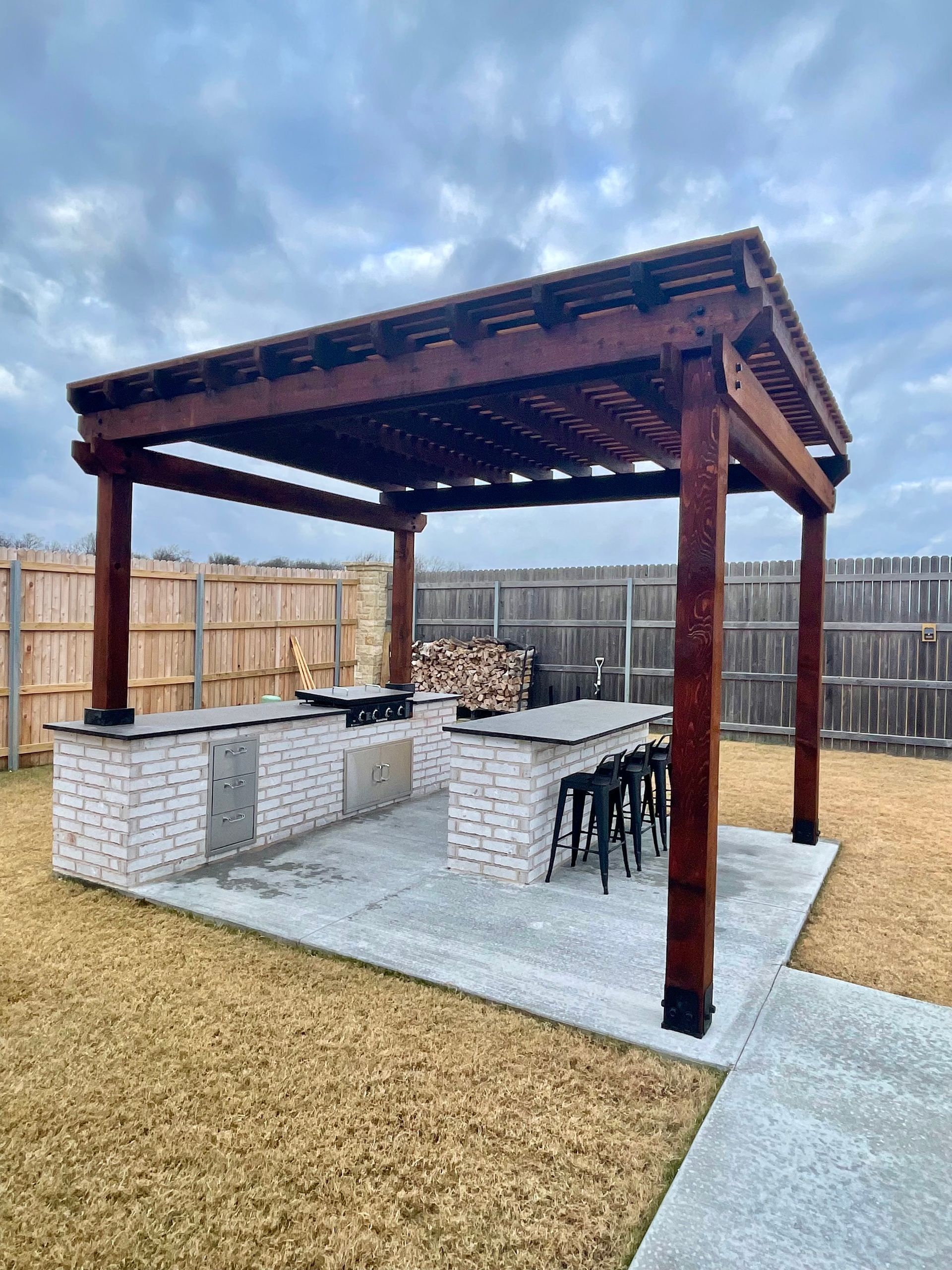 Outdoor kitchen under a brown wooden pergola with grill, countertop, and bar seating; cloudy sky.