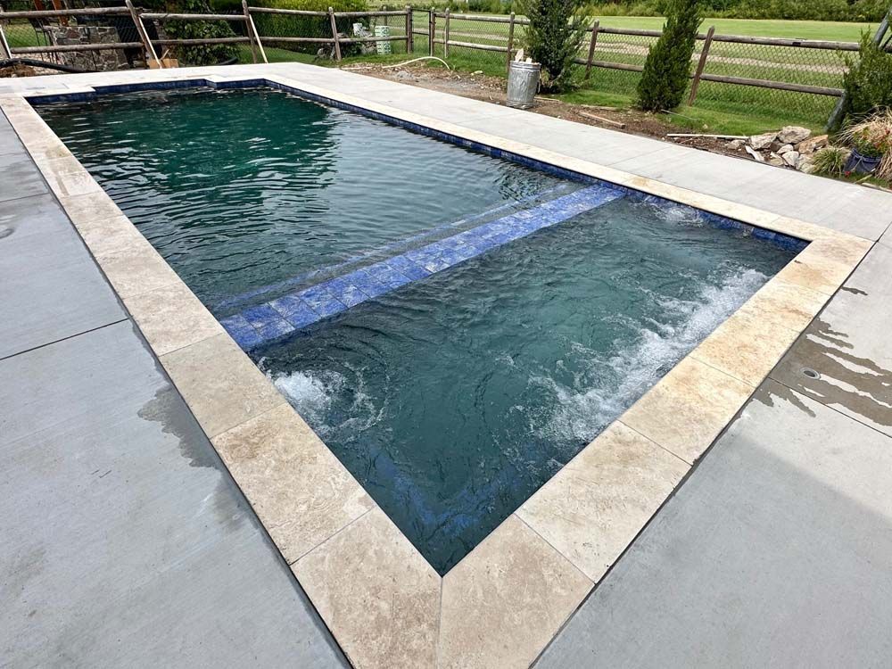 Rectangular swimming pool with tan stone border and blue tile accents. Water is clear, concrete patio surrounds it.