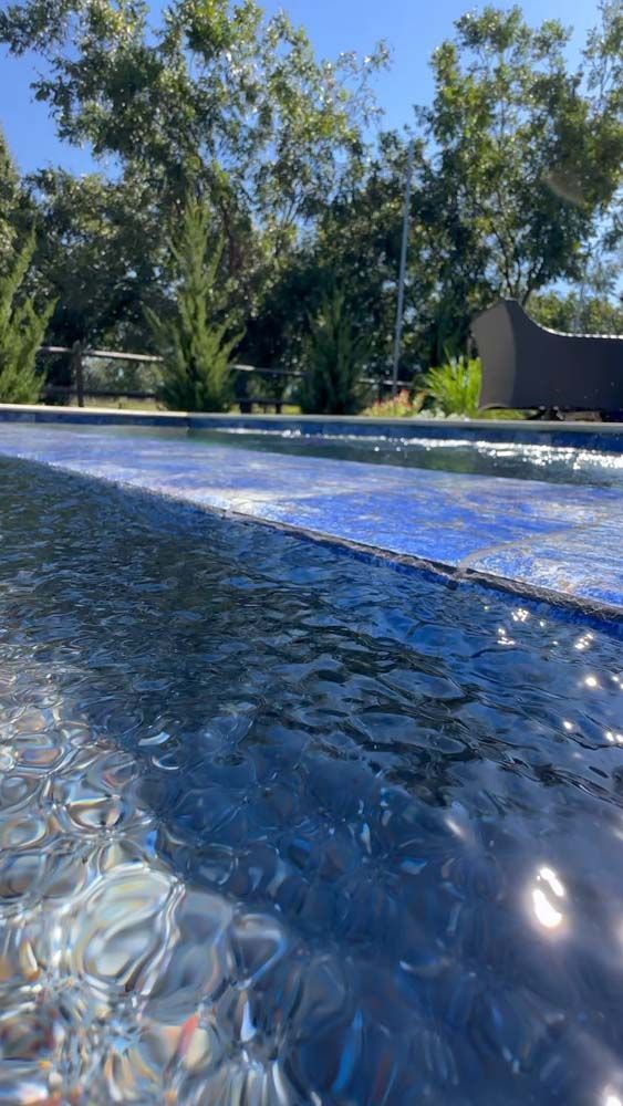 Close-up of a blue-tiled pool with shimmering water. Trees and clear blue sky in the background.