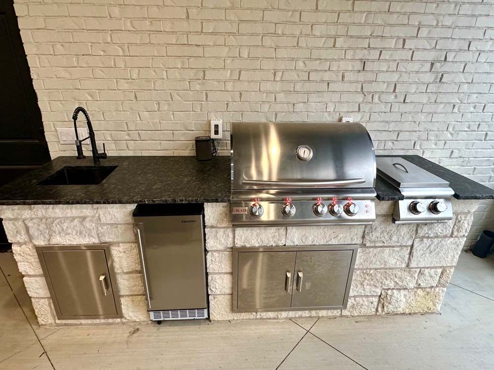 Outdoor kitchen with stainless steel grill, sink, refrigerator, set in stone-like structure.