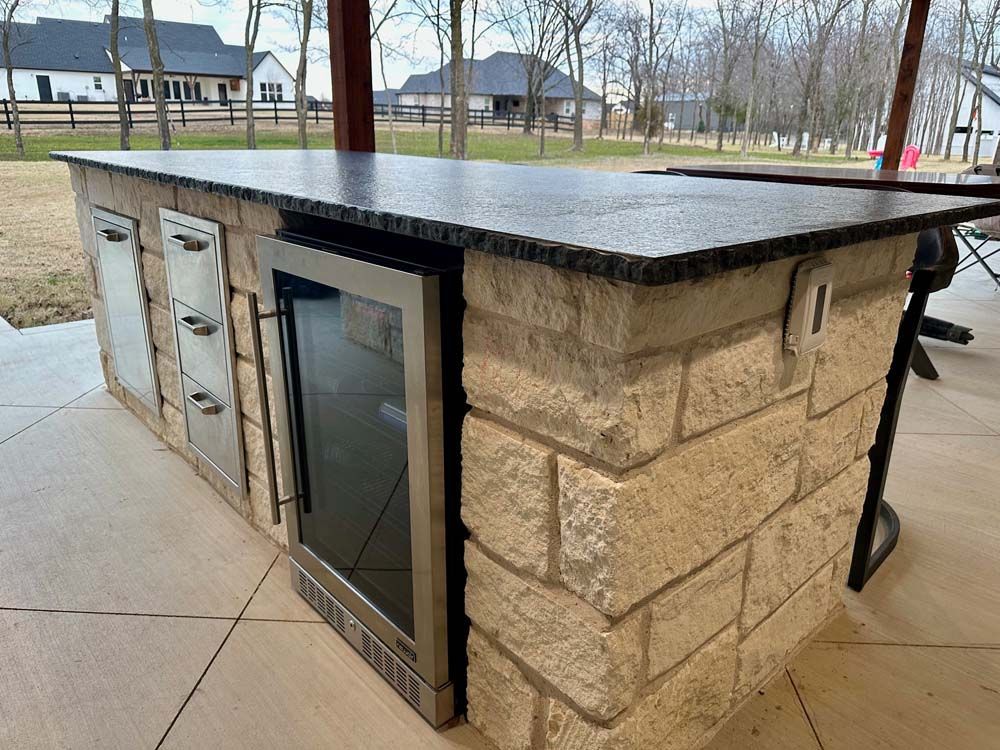 Outdoor kitchen island with a stone facade, granite countertop, refrigerator, and storage cabinets.