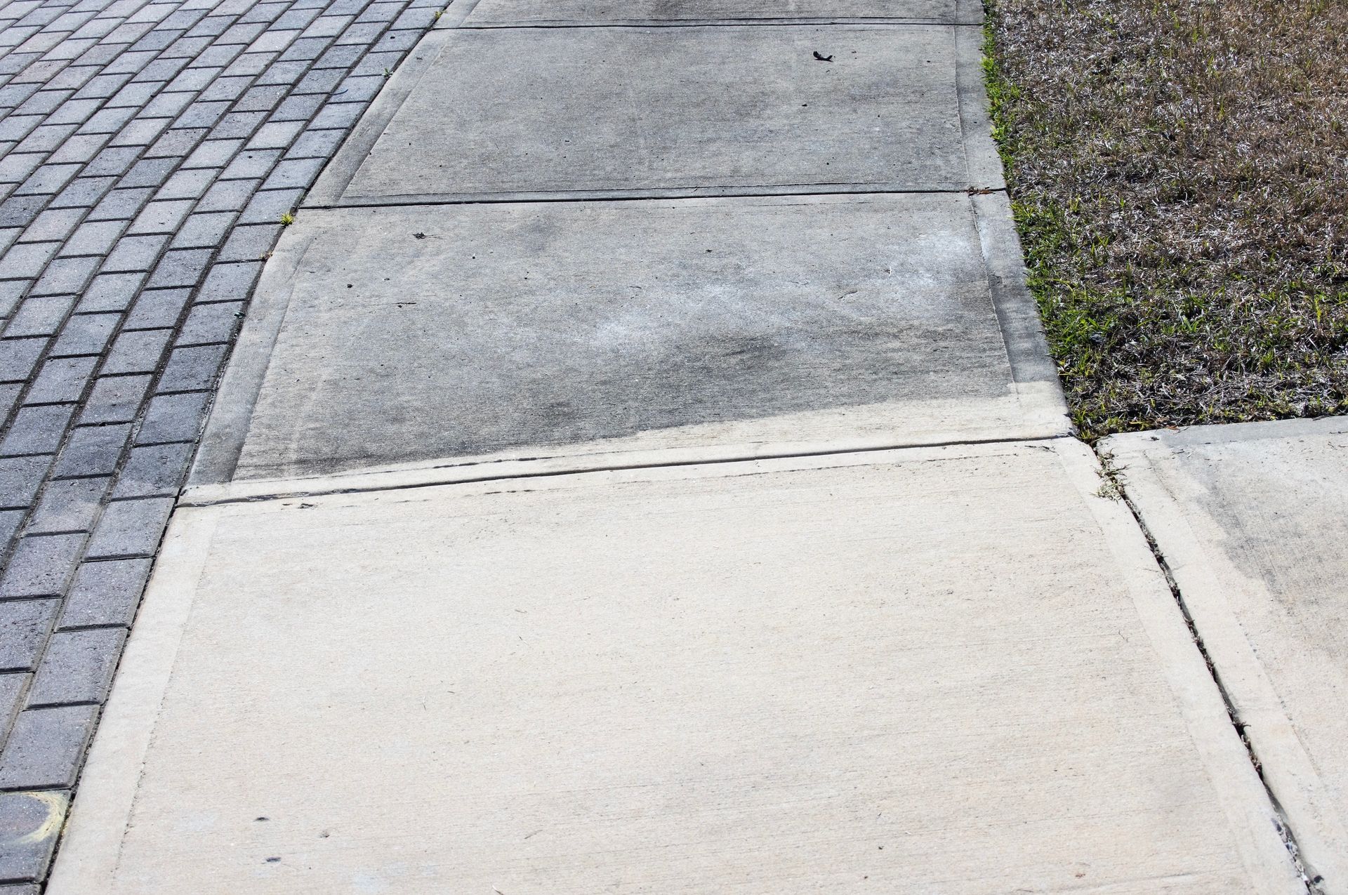 Concrete sidewalk with dark stain, next to brick pavers and dry grass.