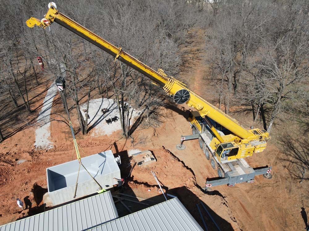 Yellow crane lifting a large gray structure onto a roof near bare trees.